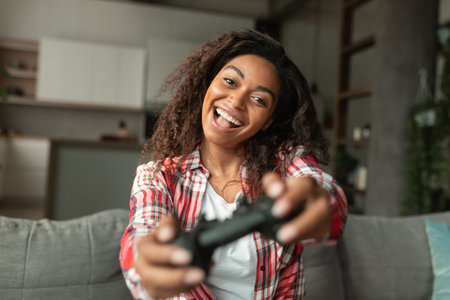 Glad excited young african american woman with open mouth playing online game with joystick, sitting...