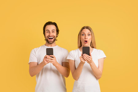 Shocked cheerful young caucasian couple in white t-shirts with open mouths chatting in smartphones