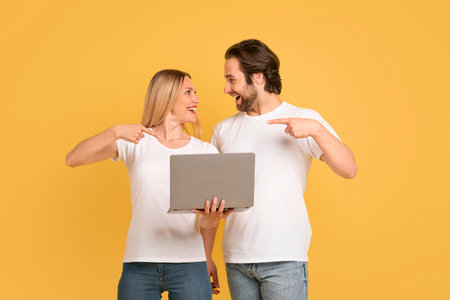Satisfied young caucasian guy and lady in white t-shirts show fingers at laptop, isolated on yellow backgroundの写真素材