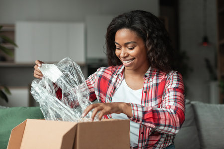 Cheerful millennial african american woman opens cardboard box and unpacks parcel in living roomの写真素材