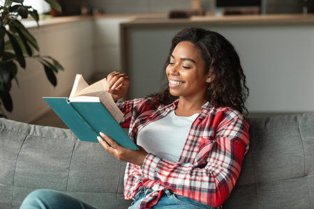Happy smart pretty millennial african american woman sitting on sofa and reading book, enjoy free timeの写真素材
