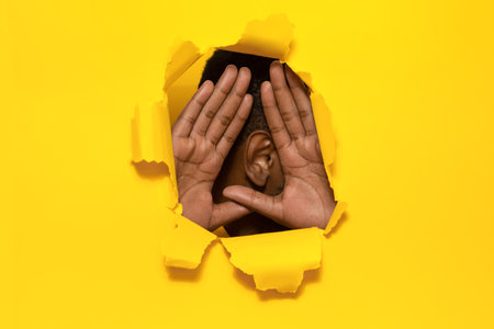 African american man holding hands near ear, listening through torn paper yellow backgroundの写真素材