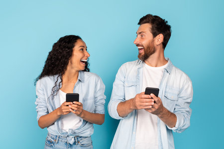 Surprised happy excited young diverse couple with smartphones look at each other, rejoice online victoryの写真素材