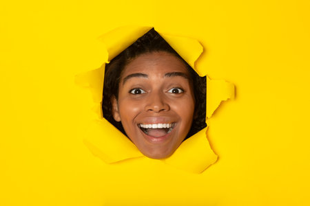 Excited African Woman Looking Through Hole In Torn Yellow Paperの写真素材