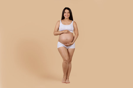 Beautiful Pregnant Female In Underwear Tenderly Embracing Belly And Smiling At Cameraの写真素材
