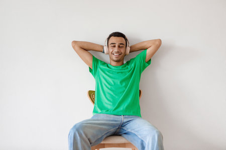 Relaxed arab man listening to music in wireless headphones, resting on chair with hands behind head over white wallの写真素材