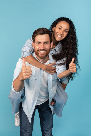 Happy millennial caucasian guy holding arab female on his back, have fun, show thumbs up, couple enjoy freedomの写真素材