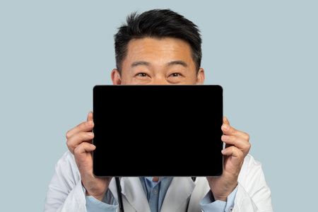 Smiling middle aged asian man worker in white coat shows tablet with blank screen