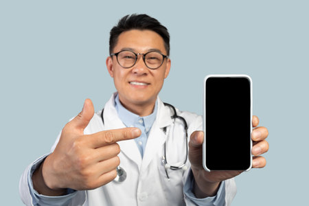 Glad middle aged chinese man therapist in white coat and glasses points on smartphone with blank screenの写真素材