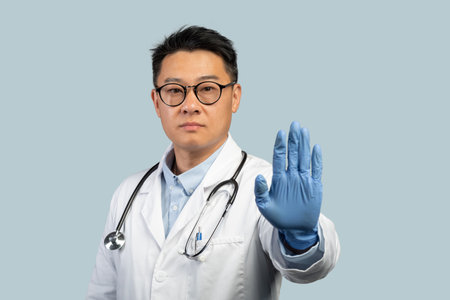 Confident serious middle aged chinese male doctor in white coat, protective gloves and glasses shows stop signの写真素材