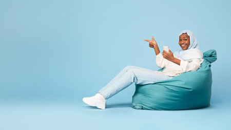 Happy african american woman sitting at bean bag with smartphone and pointing aside at copy space,...