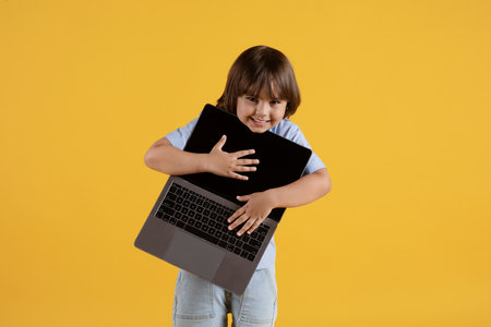 Cute tricky little boy holding open laptop and smiling to camera, orange studio background, free...
