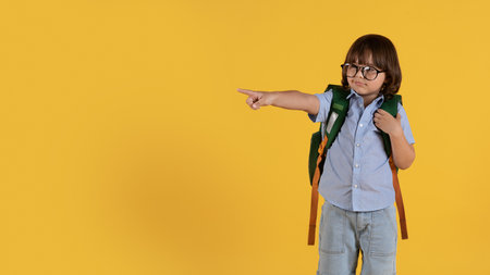Cute little boy preschooler in eyeglasses with backpack pointing aside at empty space, orange studio...