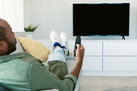 Back view of black man watching TV pointing remote control at empty plasma screen, resting on sofa at home, mockupの写真素材