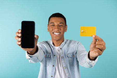 Smiling attractive millennial african american guy in jeans show smartphone with blank screen and credit cardの写真素材