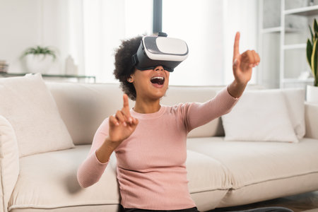 Happy surprised young african american lady with curly hair in vr glasses, touches with hands to...