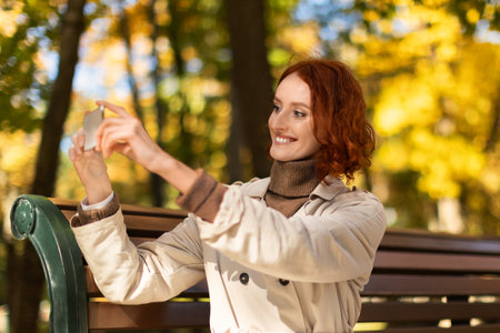 Cheerful pretty young european red-haired woman in raincoat makes photo for blog and social networksの写真素材