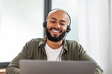 Happy latin man with beard in headphones looking at laptop and smiling, having video call from home