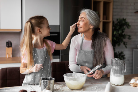 Satisfied caucasian small granddaughter and old grandmother smeared with flour, preparing dough for bakingの写真素材