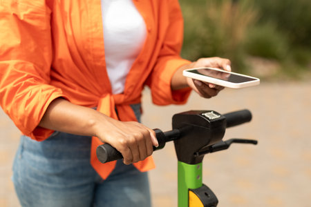 Young black female with smartphone pays money for ride on electric scooter in city park, close up, croppedの写真素材