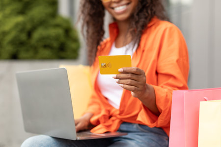 Smiling millennial black lady in casual with many bags typing on laptop, holding credit card in city, croppedの写真素材
