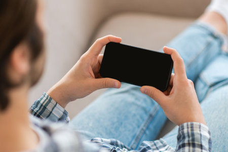 Young european male watches video, plays in game on smartphone with blank screen on sofa in living...