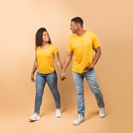 Romantic african american couple walking, holding hands, looking at each other and smiling on peach...