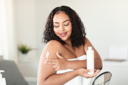 Lovely african american chubby woman wrapped in towel applying body cream on her shoulder after bath, sitting in bedroomの写真素材