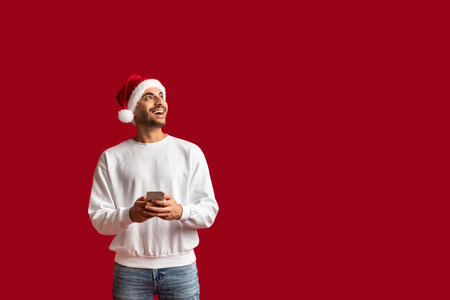 Cheerful Arab Man Wearing Santa Hat Holding Smartphone And Looking Asideの写真素材