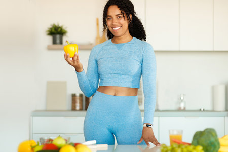 Happy african american lady in sportswear cooking fresh salad and smiling at camera, standing in kitchen interiorの写真素材