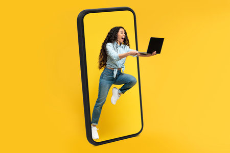 Happy excited millennial arab female with open mouth with laptop jumping out of huge smartphone screenの写真素材