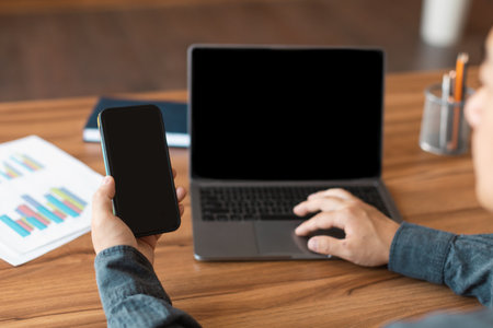 Adult chinese male looks at computer and smartphone with blank screen, works with graphs and charts...