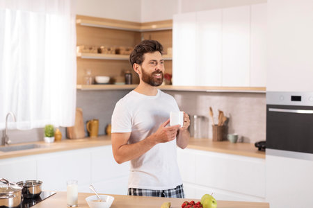 Cheerful middle aged caucasian male with beard in white t-shirt drinks hot drink looks at empty spaceの写真素材