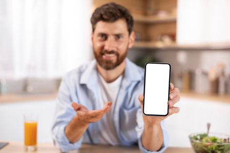 Portrait of glad adult caucasian man with beard shows phone with blank screen in kitchen interiorの写真素材