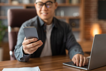 Smiling mature chinese man manager in glasses typing on smartphone at table with laptop in home...