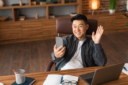 Smiling confident mature asian male waving hand at webcam phone at table with computer in officeの写真素材