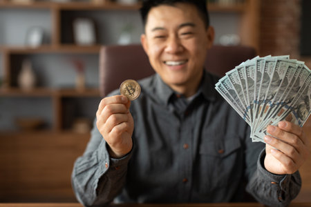 Smiling mature japanese businessman hold bitcoin coin and many dollars in office home interior, blurredの写真素材