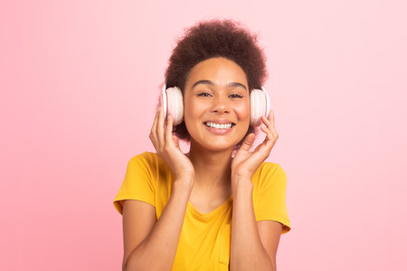 Smiling millennial african american curly female student in casual and wireless headphones, enjoy...