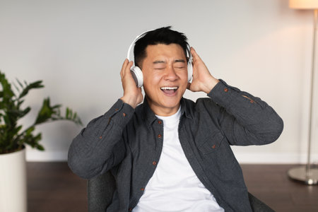Glad middle aged asian male with closed eyes in wireless headphones listens to music, enjoys free timeの写真素材