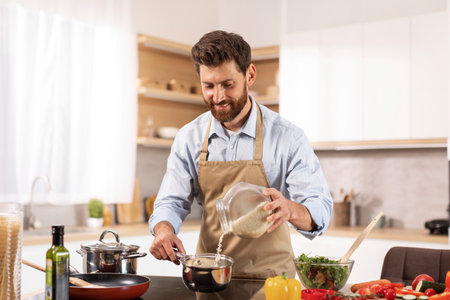 Smiling handsome adult caucasian male chef with beard in apron prepare porridge and salad in modern kitchenの写真素材