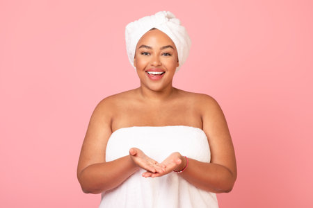 Happy black obese lady wrapped in bath towel demonstrating something on empty palms, showing invisible objectの写真素材