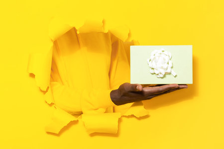 Black male hand holding or giving gift box through torn yellow paper background, closeupの写真素材