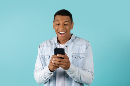 Surprised smiling young african american guy in casual typing on smartphone isolated on blue...