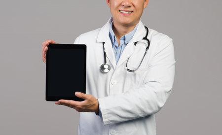 Smiling adult japanese guy therapist in white coat with stethoscope shows tablet with empty screenの写真素材