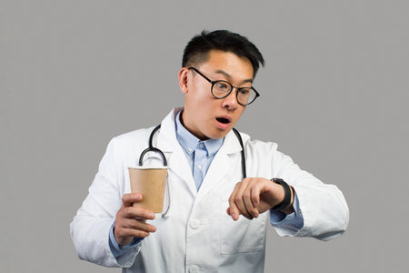 Shocked funny mature chinese male therapist in white coat, glasses with cup of coffee takeaway looks at watchの写真素材