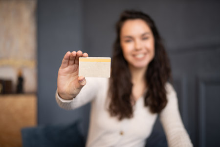 Excited young woman showing credit card and smiling at camera, sitting on sofa at home, selective focusの写真素材