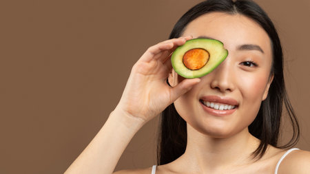 Portrait of young asian woman holding avocado hald and covering eye, smiling at camera, brown...