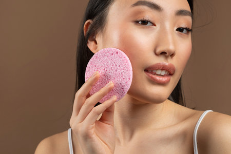 Face cleansing concept. Closeup of korean lady using soft facial cellulose sponge, washing face on brown backgroundの写真素材