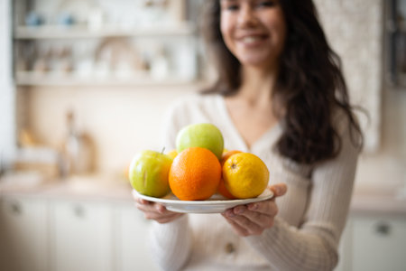 Young lady holding plate with fresh fruits apples and oranges, standing in kitchen interior, selective focus, free spaceの写真素材
