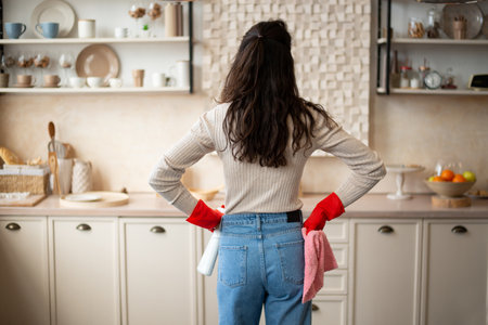 Busy young woman in rubber gloves with spray and rag in hands looking at cleaning result in kitchen interior, back viewの写真素材